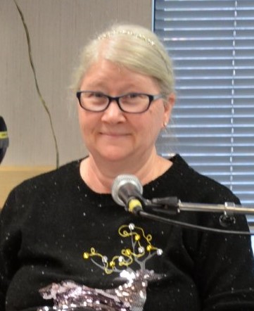 A silver-haired woman wearing eye glasses and a black sweater with a sequined reindeer smiles at the audience. A microphone is in front of her.